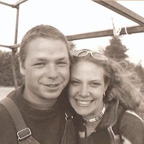 An older black and white photo of a young Mike and Fiona standing together in front of the canoe racks of a truck.