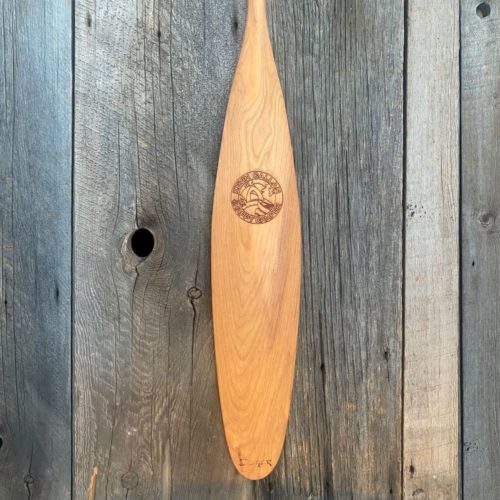 A detailed view of a cherry Woodland canoe paddle blade with a laser engraved Kevin Callan-Happy Camper logo on the blade resting on a weathered barn board background.