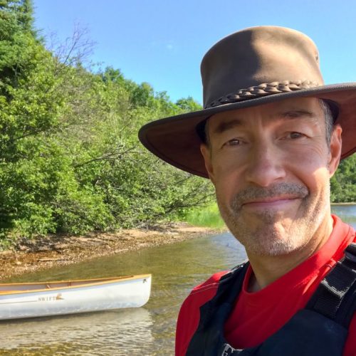 Selfie photo of (SwiftyPaddler) a man in hat wearing PFD, standing in front of a canoe on a forested shoreline.