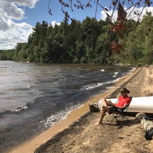Man sitting in a camp chair, reading a book, on a forested beach beside a canoe somewhere in Ontario