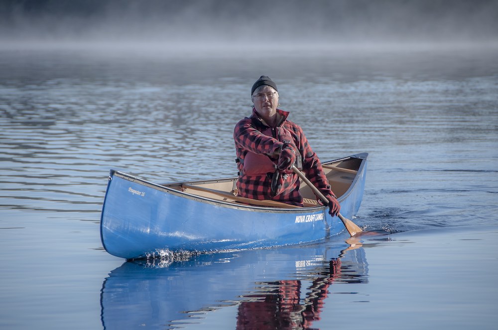 Kevin Callan, The Happy Camper, paddles a blue Nova Craft Prospector canoe on a misty blue lake. He is wearing a buffalo plaid coat and red PFD.