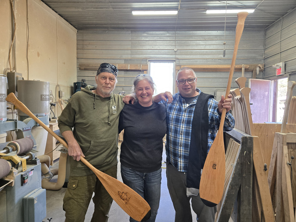 Hap Wilson stands to the left with his arm around his wife, Andrea Wilson, who is flanked by Mike on the right. Both men are holding canoe paddles. They are standing in shop with some machines and paddles in the background.